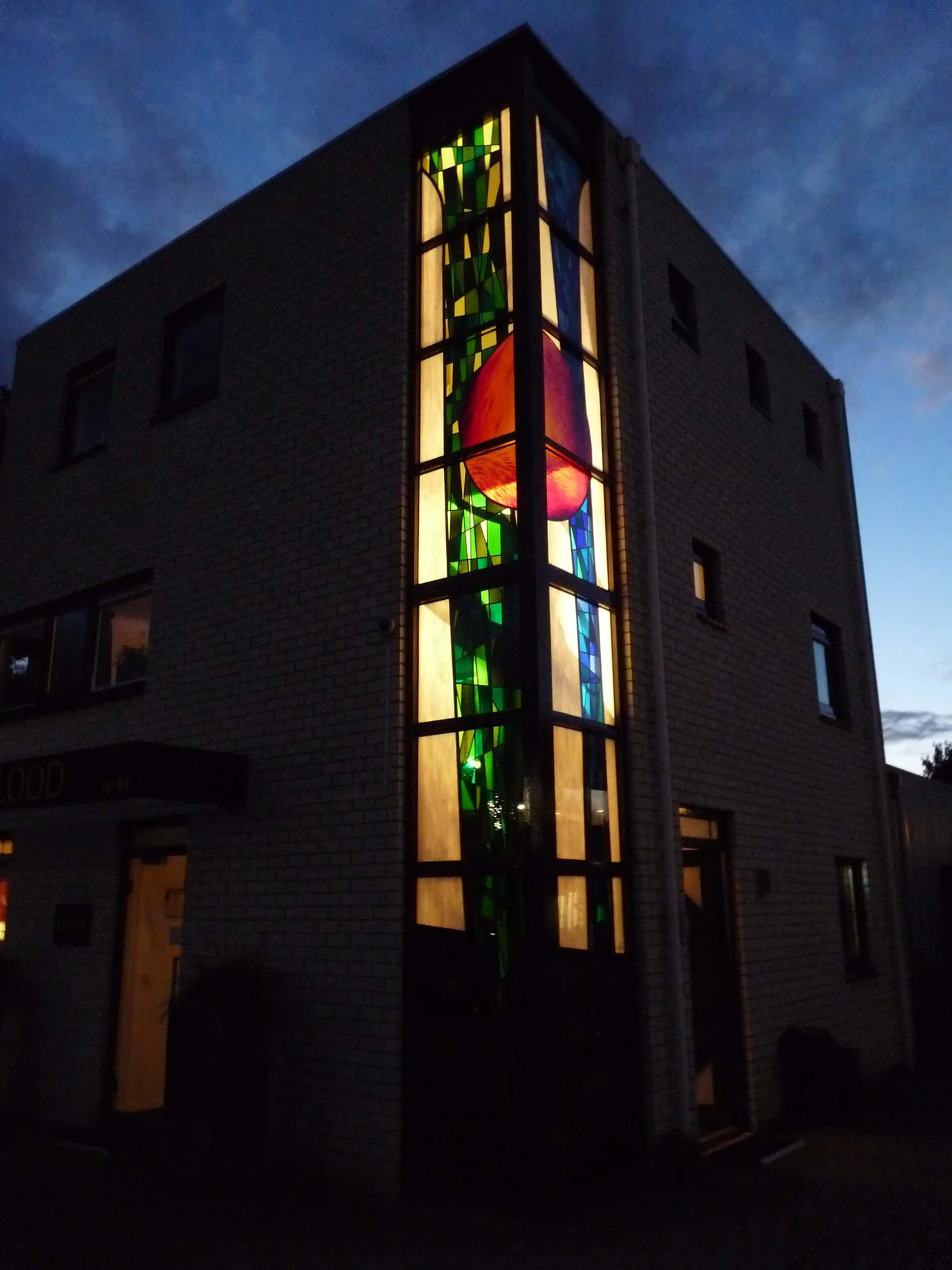 Het bedrijfspand aan Kerkenbos bij avond, met het hoge hoekraam van Ted Felen van binnenuit verlicht in groen, blauw, amber en een groot rood vlak